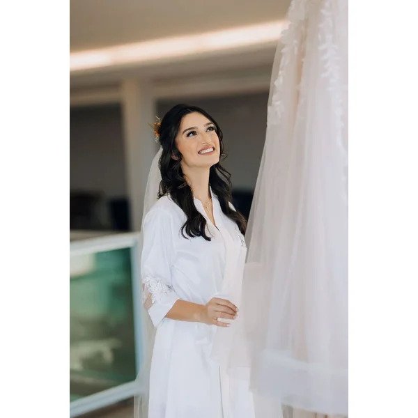 noiva com robe longo olhando para vestido de noiva pendurado