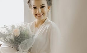 noiva sorrindo com robe branco segurando buque envolto por uma redinha de protecao branca