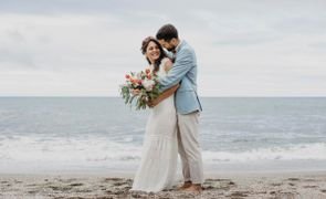 marido de terno azul claro e noiva com buque grande posando na praia