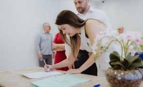 noiva assinando documento que esta sobre a mesa com o marido ao lado vaso de flor sobre a mesa e pessoas atras