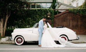 noivo beijando testa da noiva com carro antigo branco ao fundo