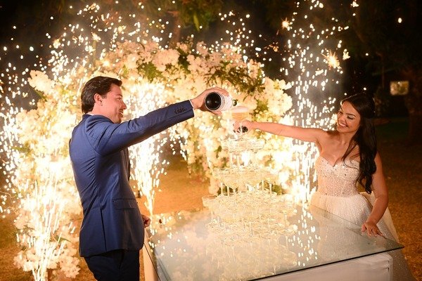 marido e esposa despejando champanhe em torre de tacas com flores e fogos ao fundo