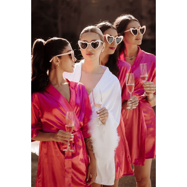 noiva com robe branco e tres madrinhas com robe pink todas com oculos de coracao e segurando taca de champanhe
