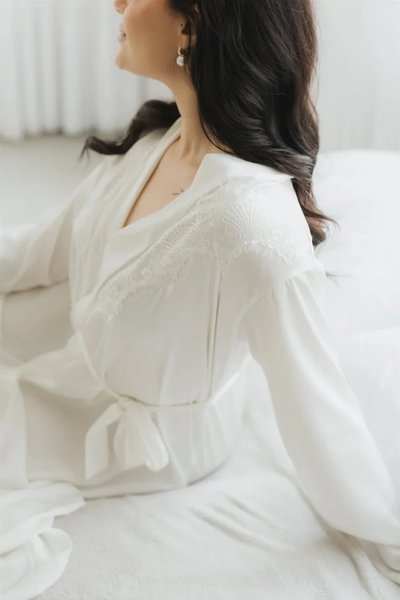 modelo de lado sentada na cama usando robe noiva longo com aplicacoes de renda no peitos e nas mangas robe penelope