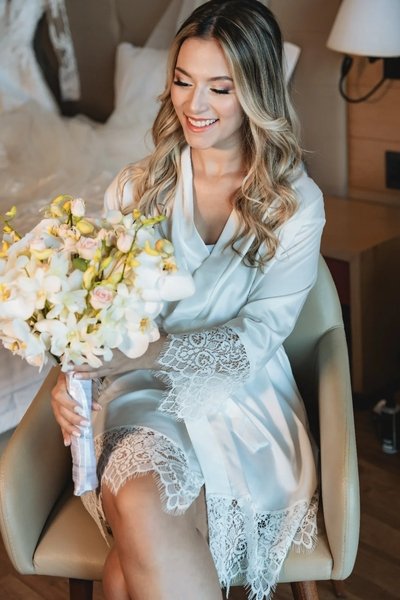 noiva sentada em cadeira segurando buque usando robe personalizado para noivas com renda nas barras robe ana carolina