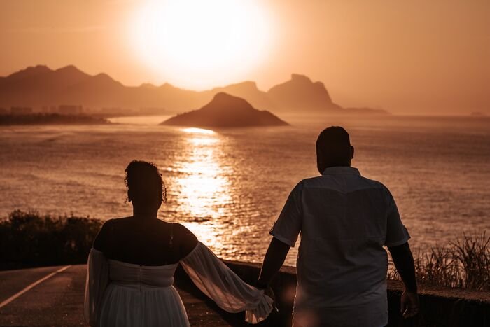 casal de noivos de mao dada de costas olhando para o mar e mobtes totalmente ensolarado