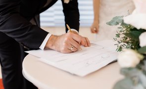 foco em maos do noivo assinando documento com rosas brancas sobre a mesa e noiva ao fundo